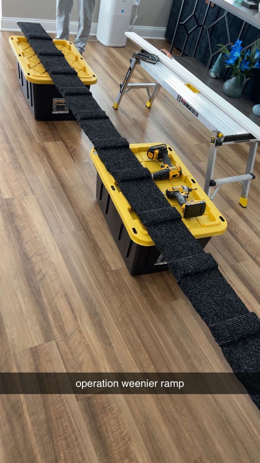 Ramp bottom board with carpet-covered cleats laid across two yellow-lid storage totes indoors, drills resting on a tote and a work platform beside it, captioned operation weenier ramp